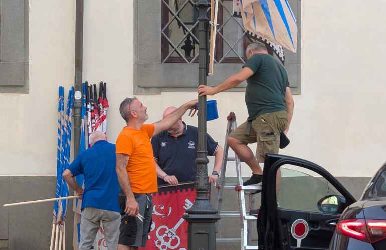 Volontari della Lega dei Rioni allestiscono le bandiere del Palio sui lampioni di piazza Mazzini a Pescia