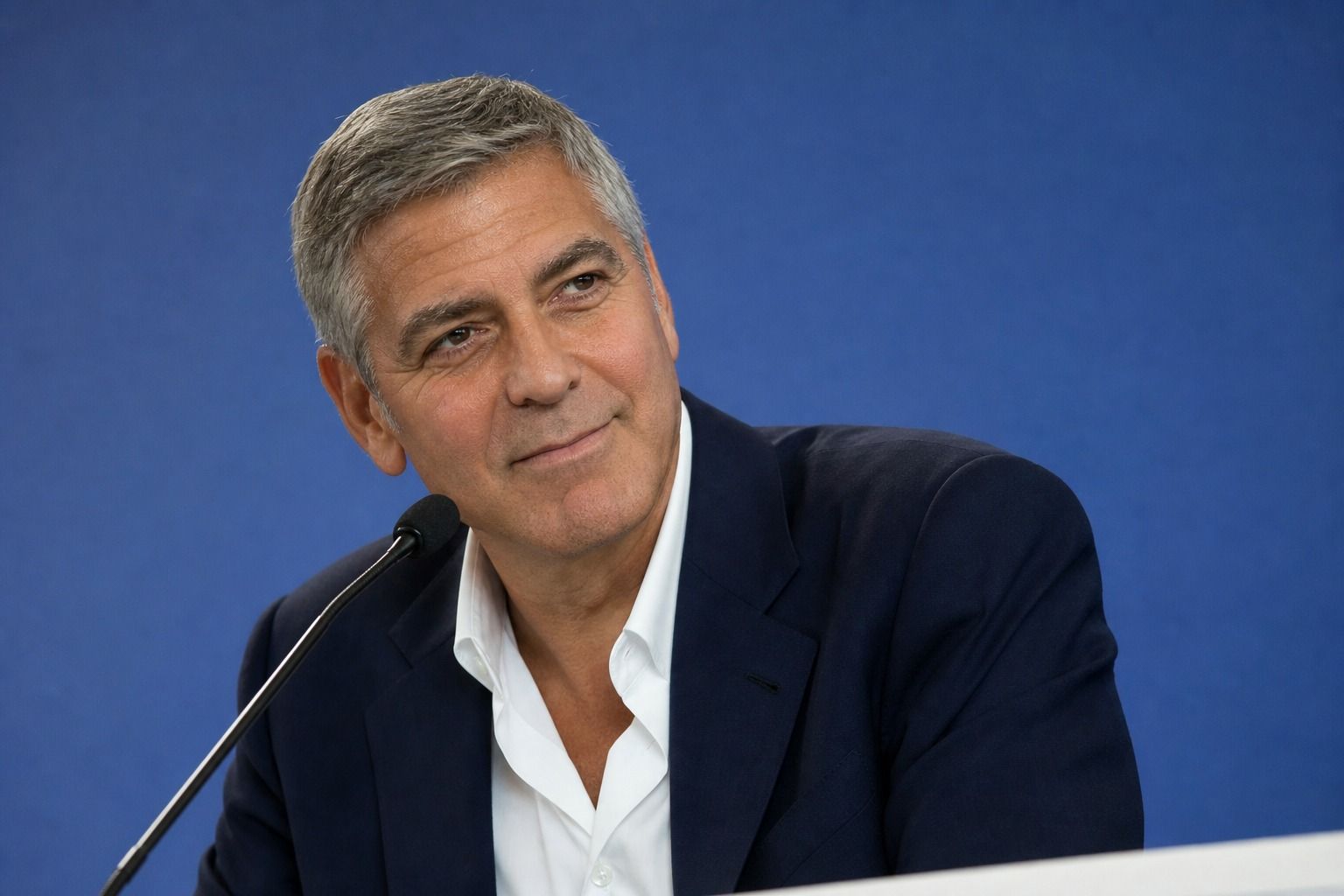 George Clooney in giacca blu a una conferenza stampa a Venezia, microfono in primo piano